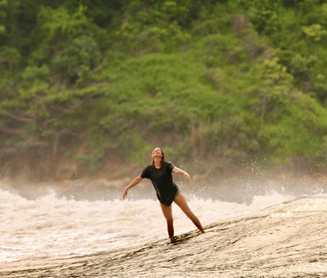 Catherine standing in the surf with her arms open, laughing up at the sky as spray rises behind her